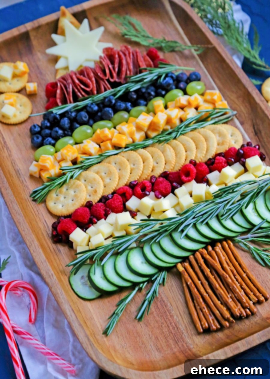 A top-down view of a partially completed Christmas Tree Charcuterie Board, showing an organized arrangement of cheeses, salami, blueberries, raspberries, and pretzel sticks on a light wooden background.