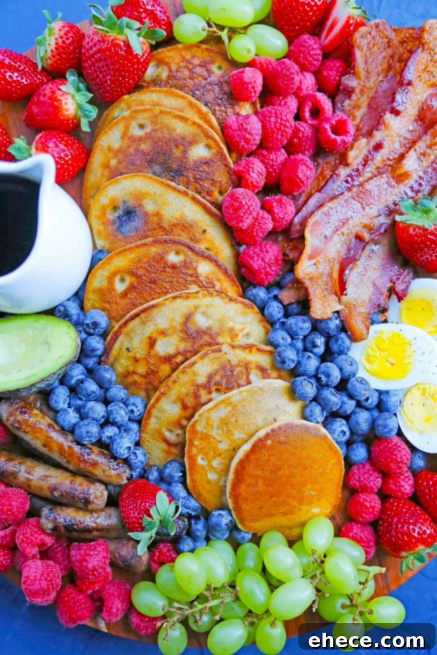 An overhead shot of a vibrant breakfast board featuring a variety of hot and cold items, including eggs, bacon, pancakes, fresh berries, and grapes.