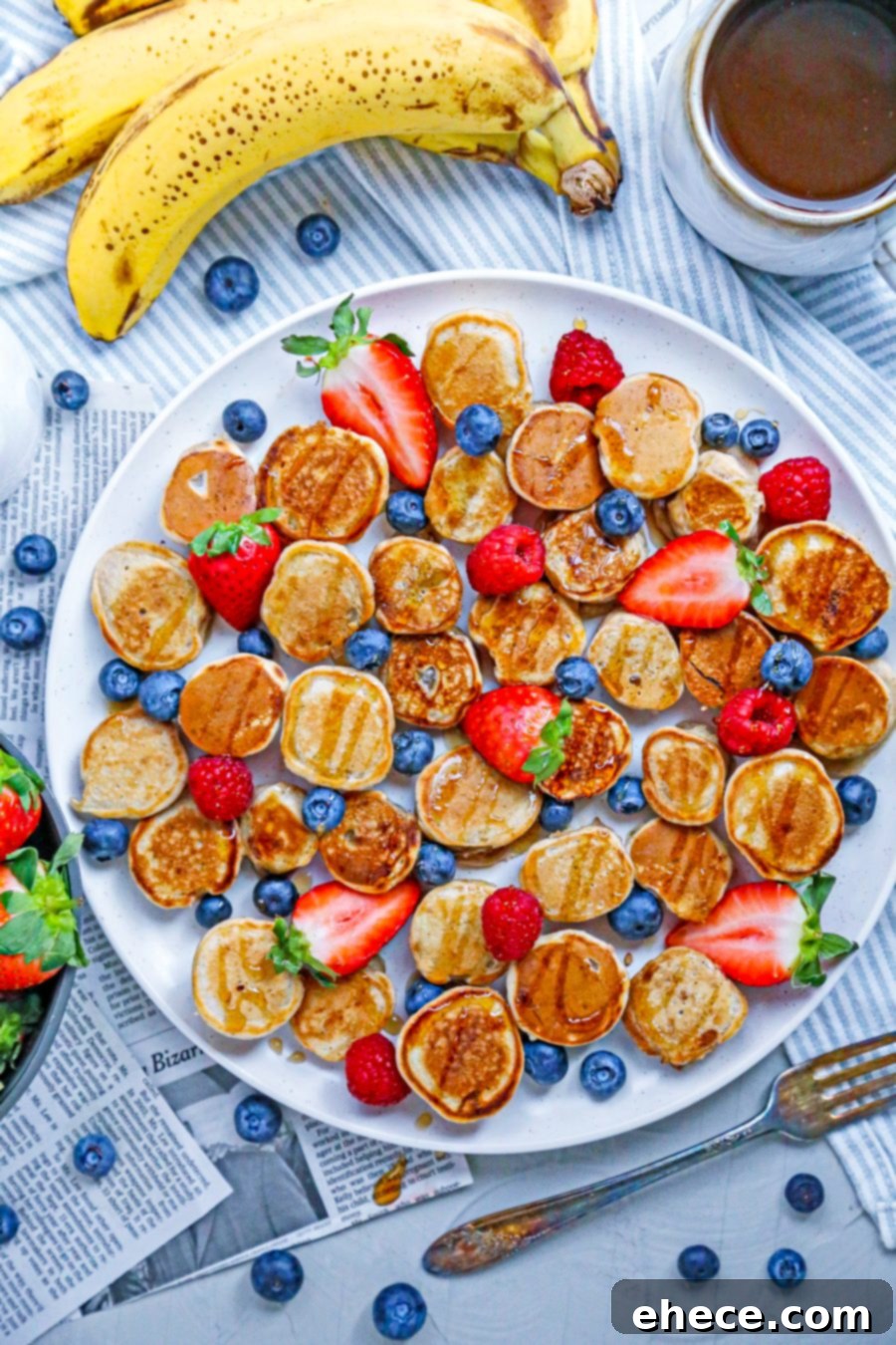 A top-down view of a plate filled with mini banana pancake bites, fresh berries, and a small pitcher of maple syrup